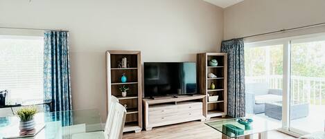 A modern living room with a glass dining table, two ceiling fans, a flat-screen TV, and a sofa.
