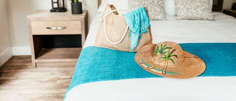 Una cama bien hecha con una colcha azul, un sombrero de mimbre y un bolso color beige sobre la cama. Se ve un mesita de noche con una lámpara y una planta pequeña al lado de la cama.