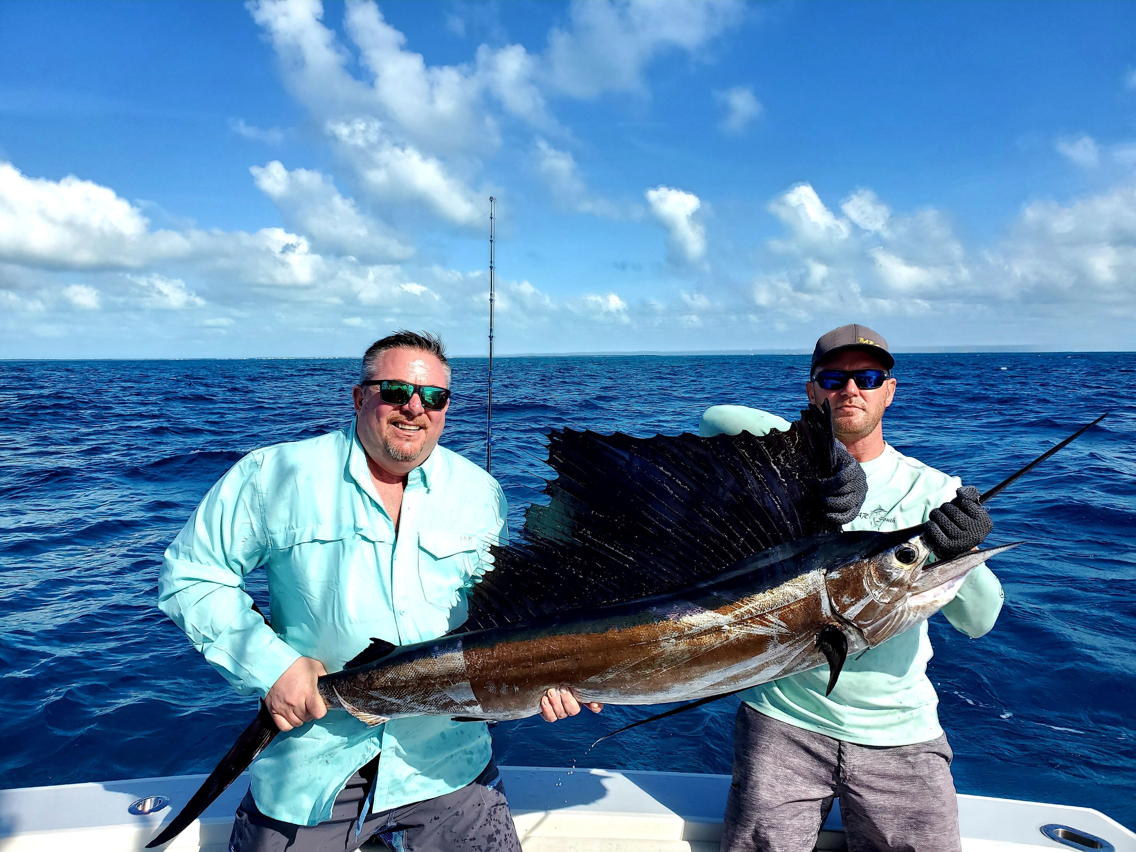 Two people holding a large fish on a boat with a clear blue sky and ocean in the background.