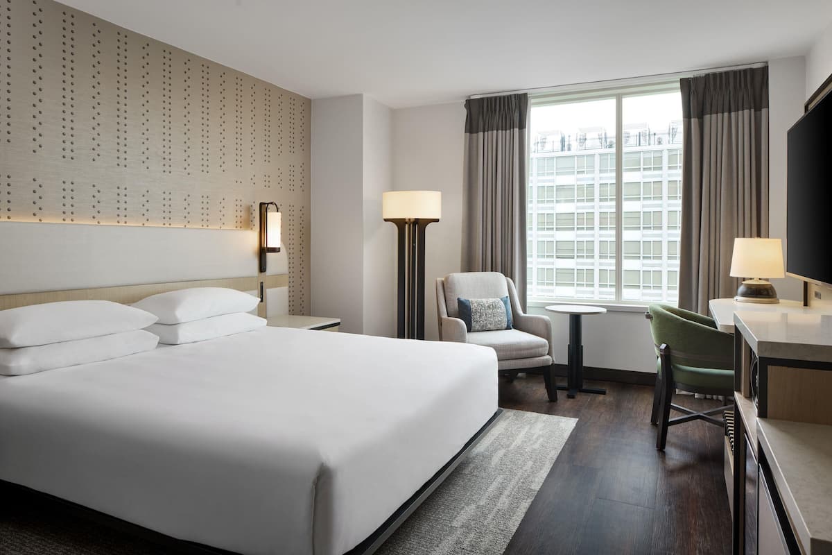 Ein Hotelzimmer mit einem großen Bett, einem Schreibtisch, einem Sessel und einer Lampe, mit Blick auf ein Gebäude.