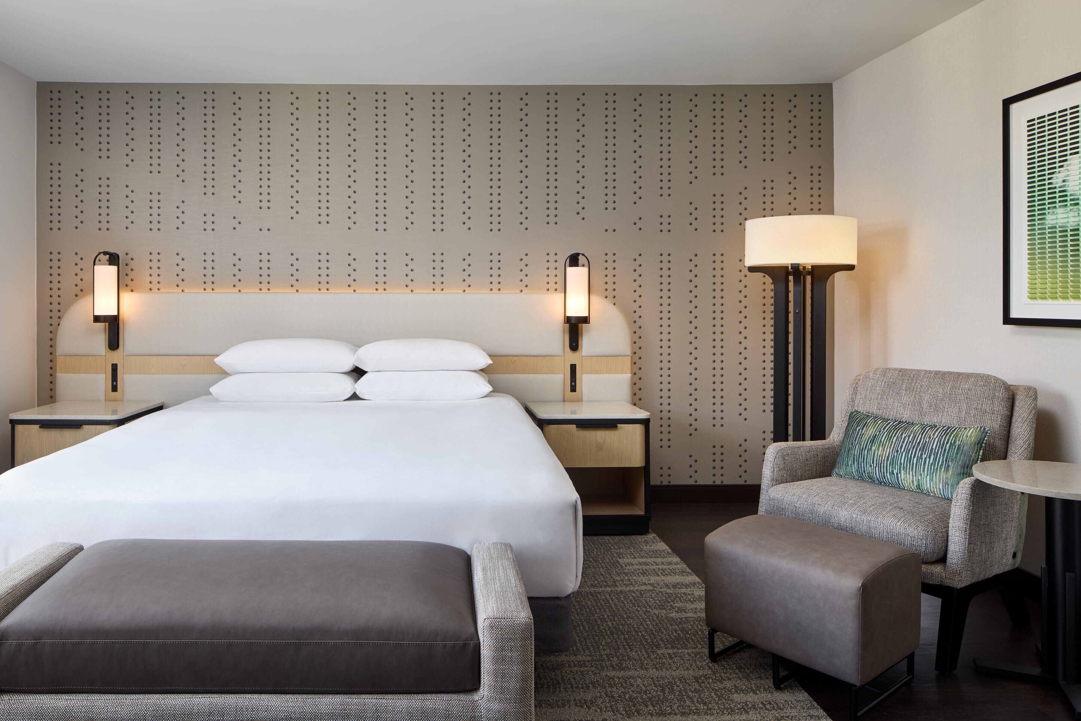 A hotel room with a large bed, two bedside tables with lamps, a chair, a small table, and a patterned wall.