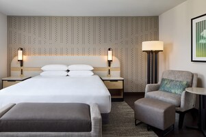 A hotel room with a large bed, two bedside tables with lamps, a chair, a small table, and a patterned wall.