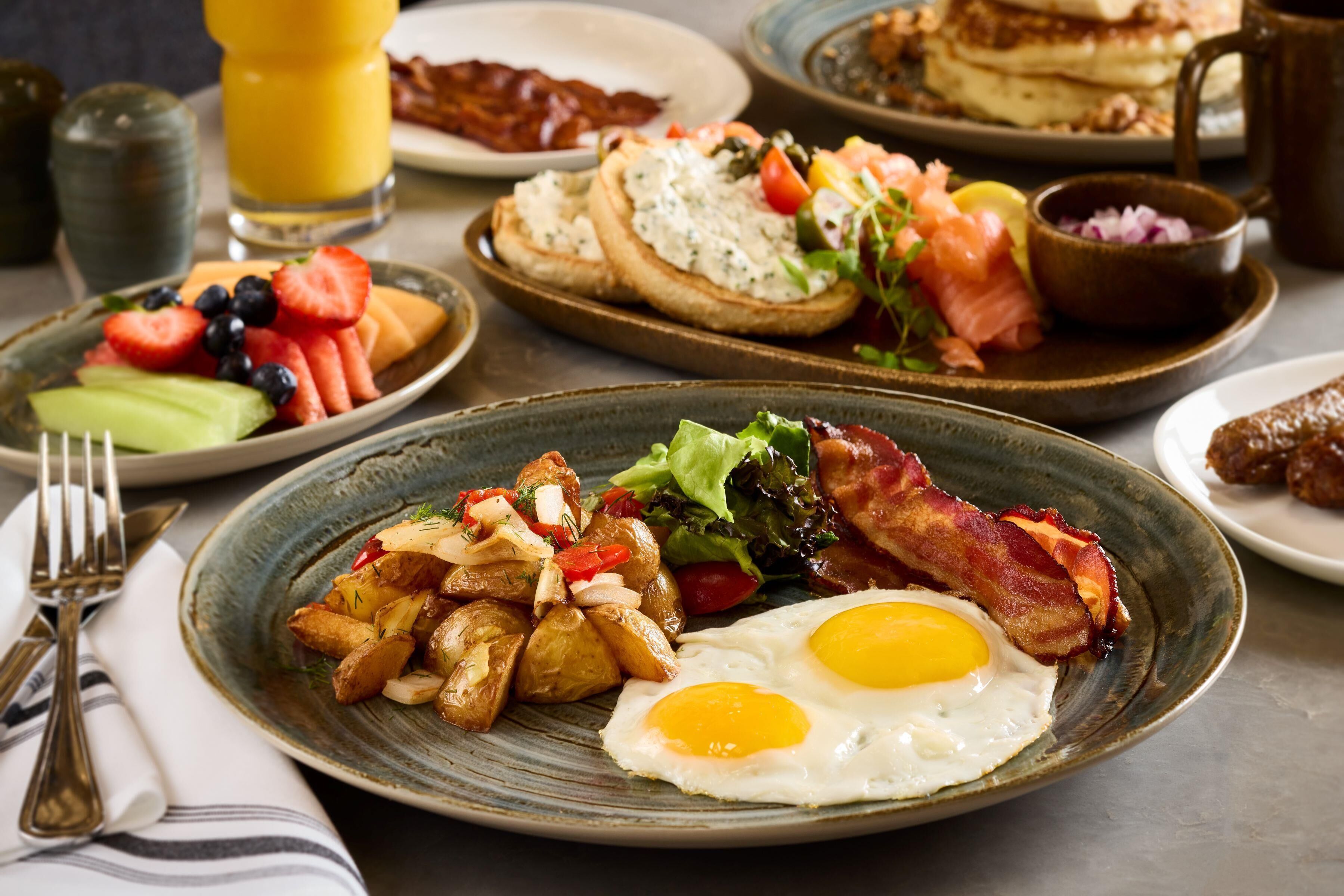 A breakfast spread with eggs, bacon, potatoes, and various fruits on plates.