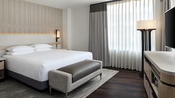 A modern hotel room with a large bed, a bench, a desk, a flat-screen TV, and a wall-mounted light fixture.