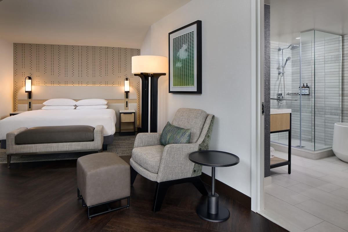 Ein Hotelzimmer mit einem Bett, einem Sessel, einem kleinen Tisch, einer Lampe, einem gerahmten Bild und einer Dusche.