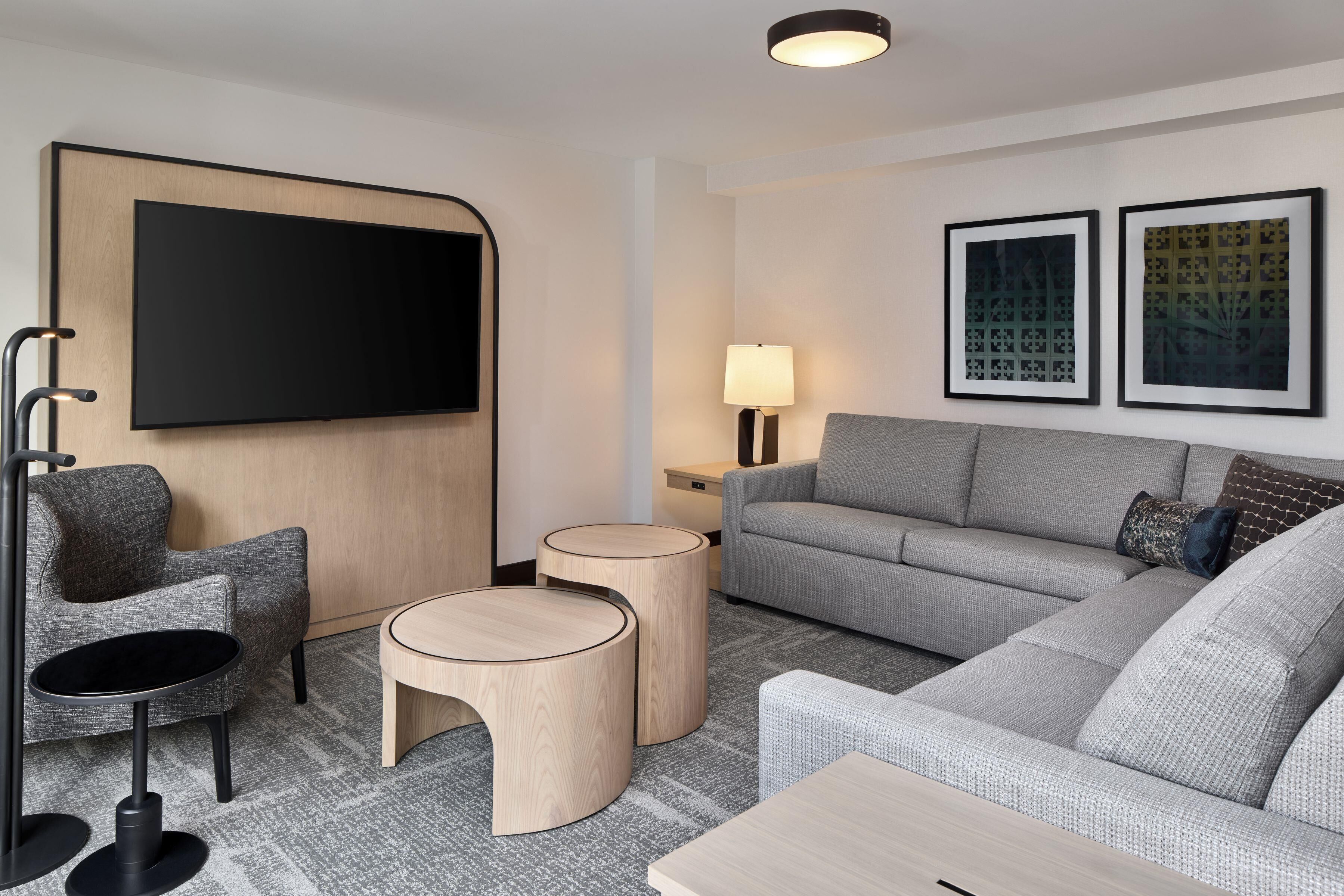 A modern hotel room with a grey sofa, a flat-screen TV mounted on a wooden panel, two round wooden side tables, a lamp, and two framed pictures on the wall.