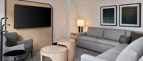 Quarto de hotel moderno com sofá cinzento, televisão de tela plana fixada em um painel de madeira, duas mesinhas de cabeceira redondas de madeira, um abajur e duas quadros emoldurados na parede.