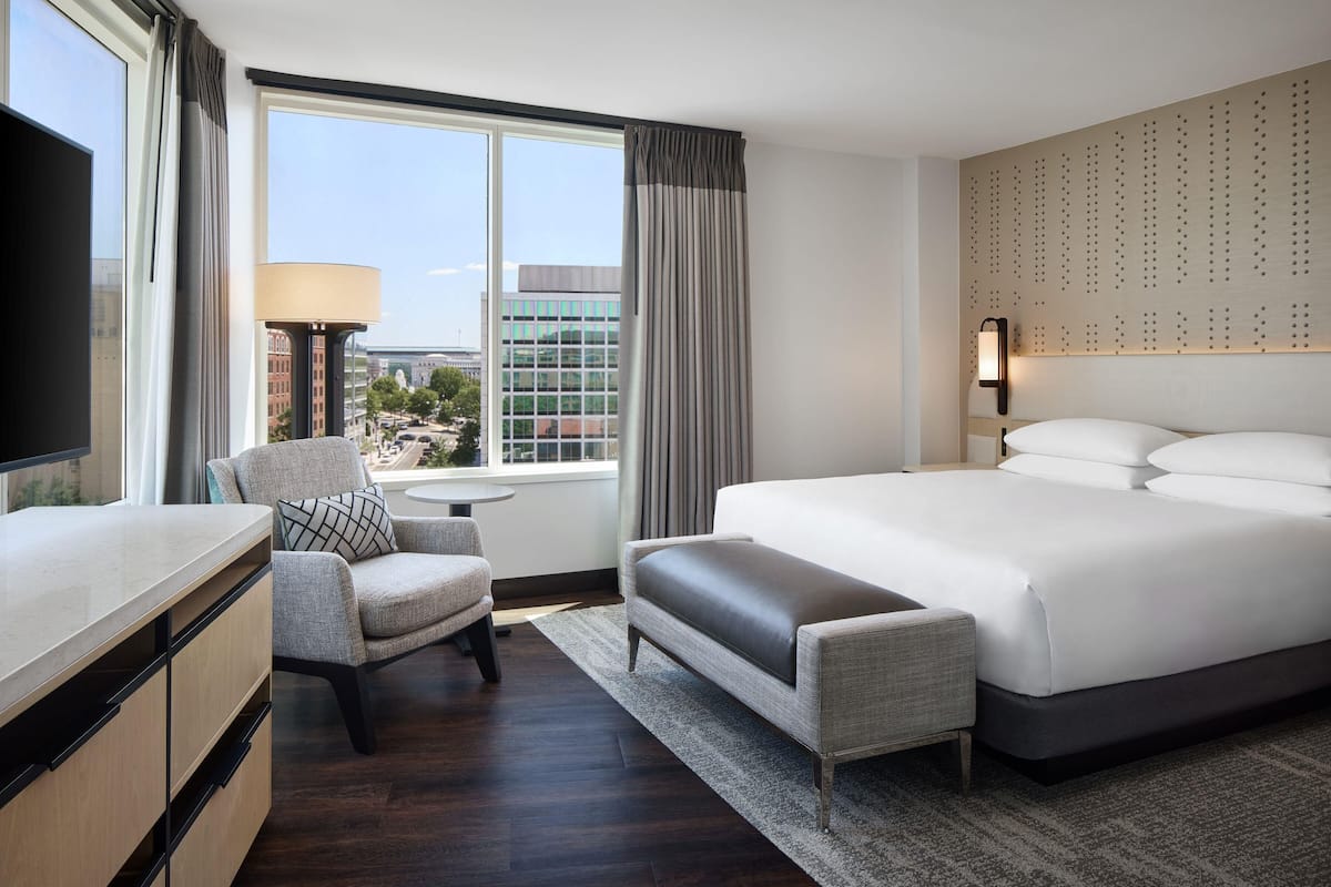 Ein Hotelzimmer mit einem großen Bett, einem Schreibtisch, einem Sessel, einem Fernseher, einem Fenster mit Vorhängen und Blick auf die Stadt.