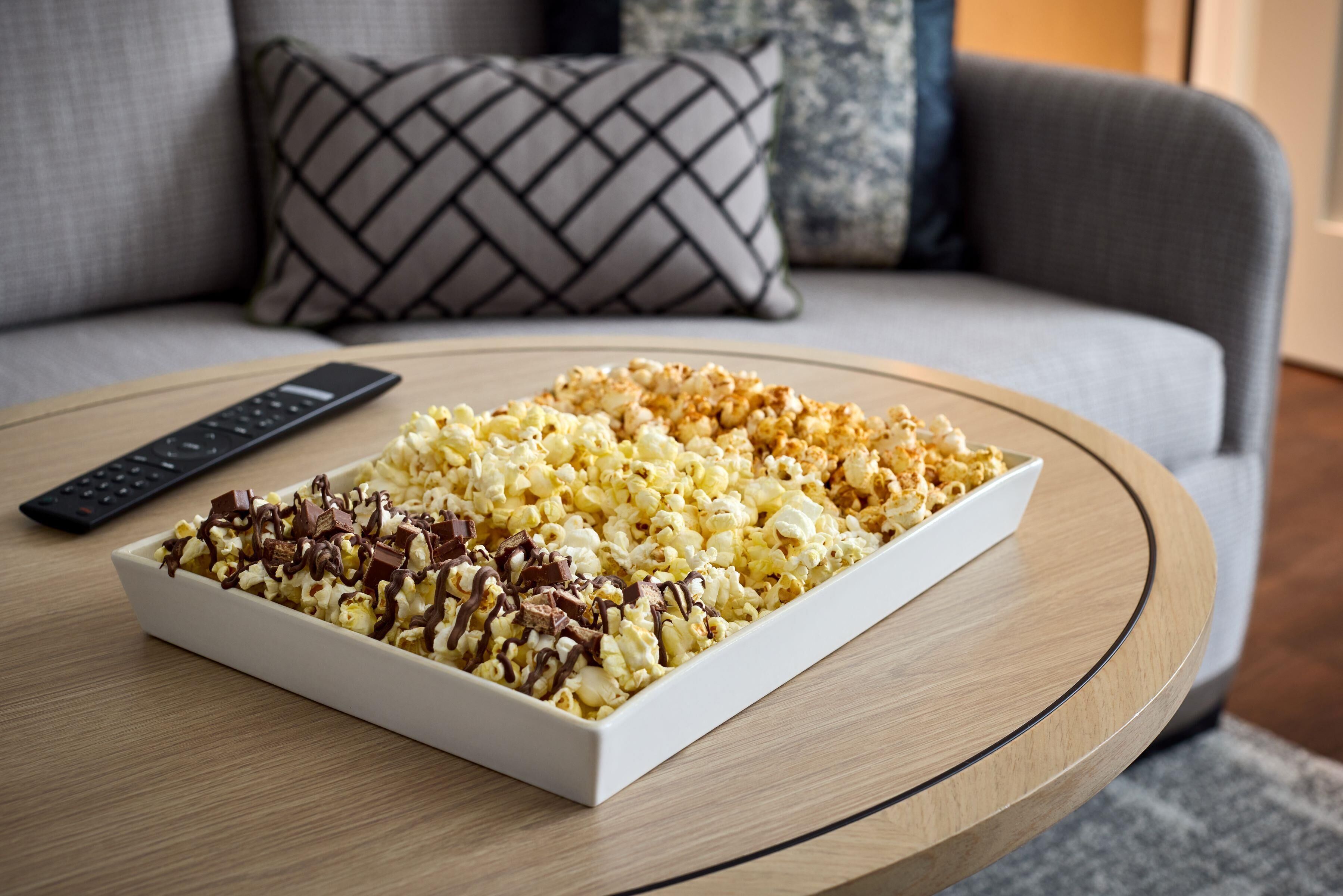 A white rectangular bowl of popcorn on a round wooden table with a remote control beside it.