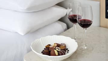 A bed with white linens, a bowl of assorted chocolates, and two glasses of red wine on a marble surface.