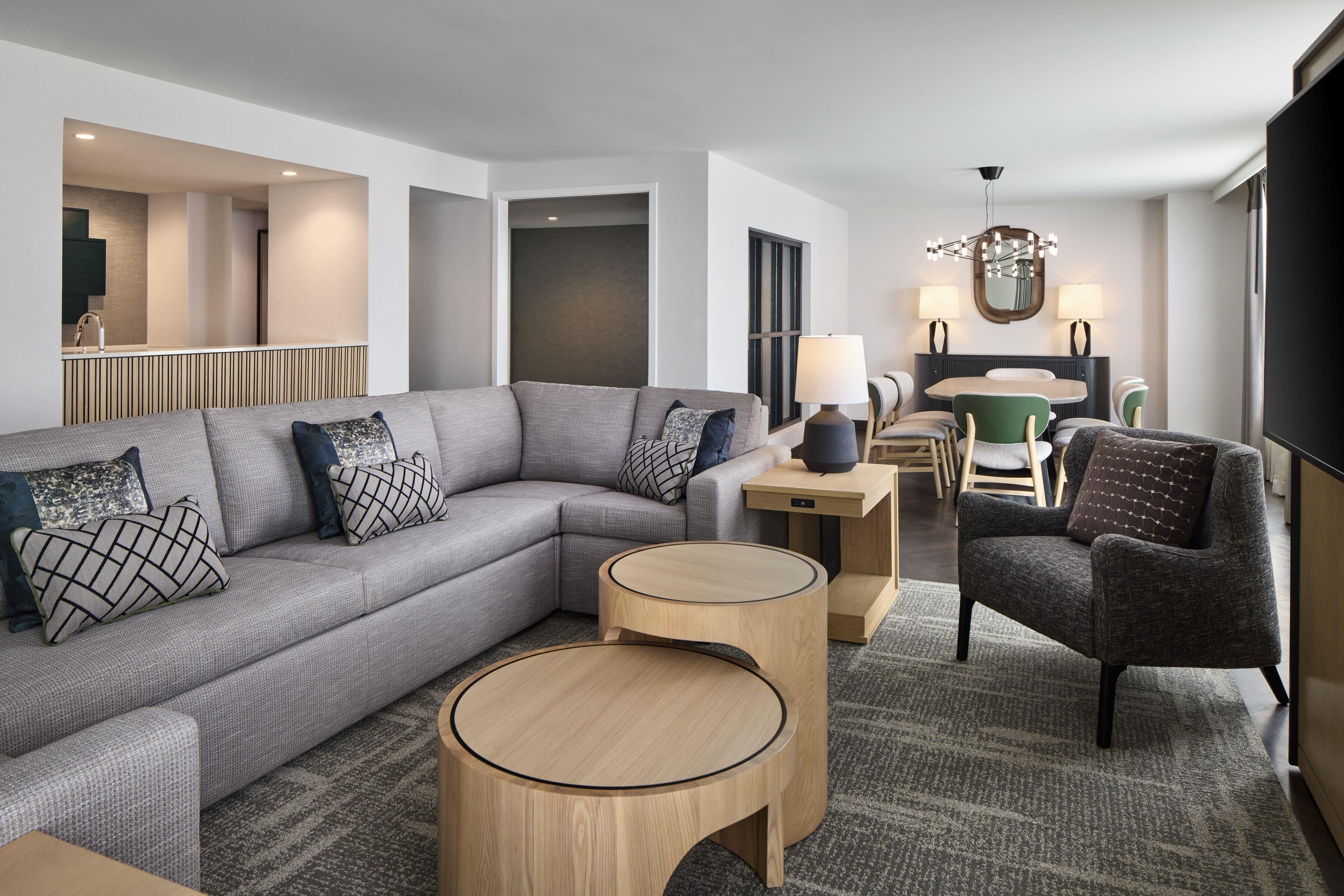 A modern living room with a grey sectional sofa, round wooden side tables, a green armchair, and a dining area with a chandelier.