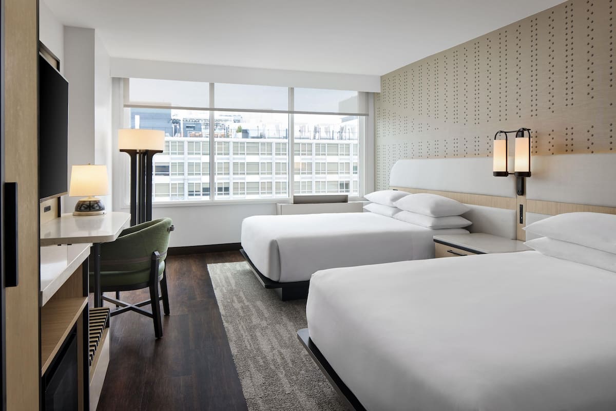 Ein Hotelzimmer mit zwei Betten, einem Schreibtisch, einem Stuhl, einem Fernseher und einem Fenster mit Blick auf die Stadt.