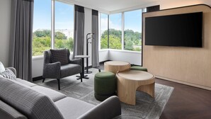 Una habitación de hotel moderna con un ventanal grande, un televisor de pantalla plana, un sofá, una silla y dos otomanas.