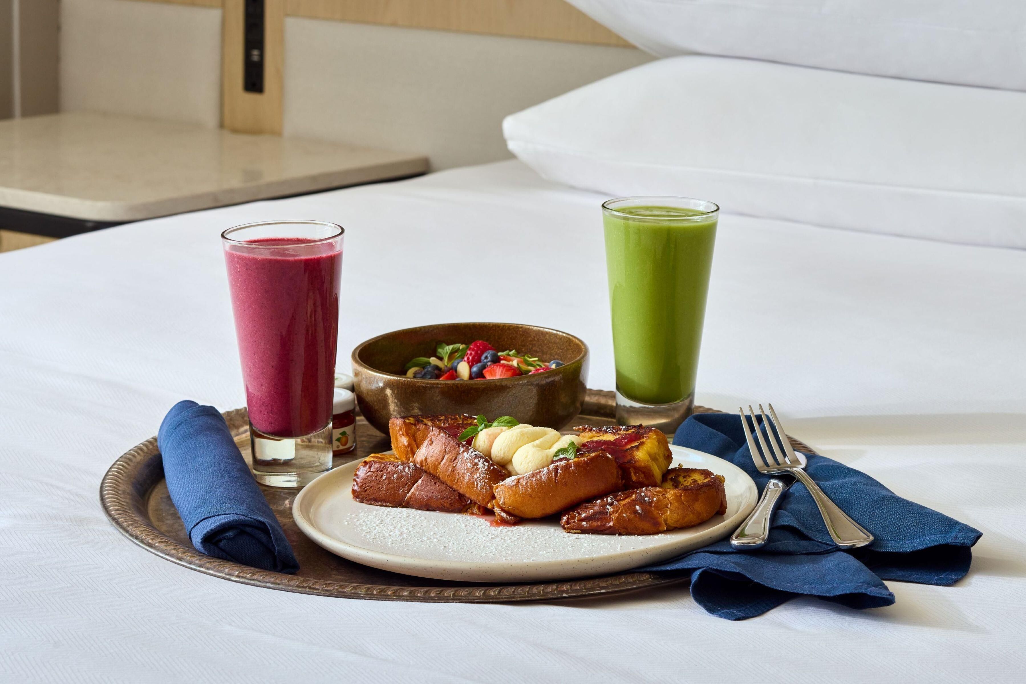A breakfast tray with pancakes, a bowl of fruit salad, and two glasses of juice on a bed.
