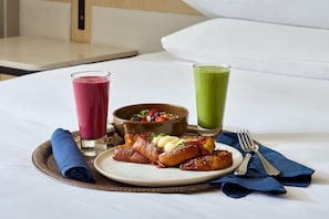 Bandeja de desayuno con panqueques, ensalada de frutas y dos vasos de jugo sobre una cama.