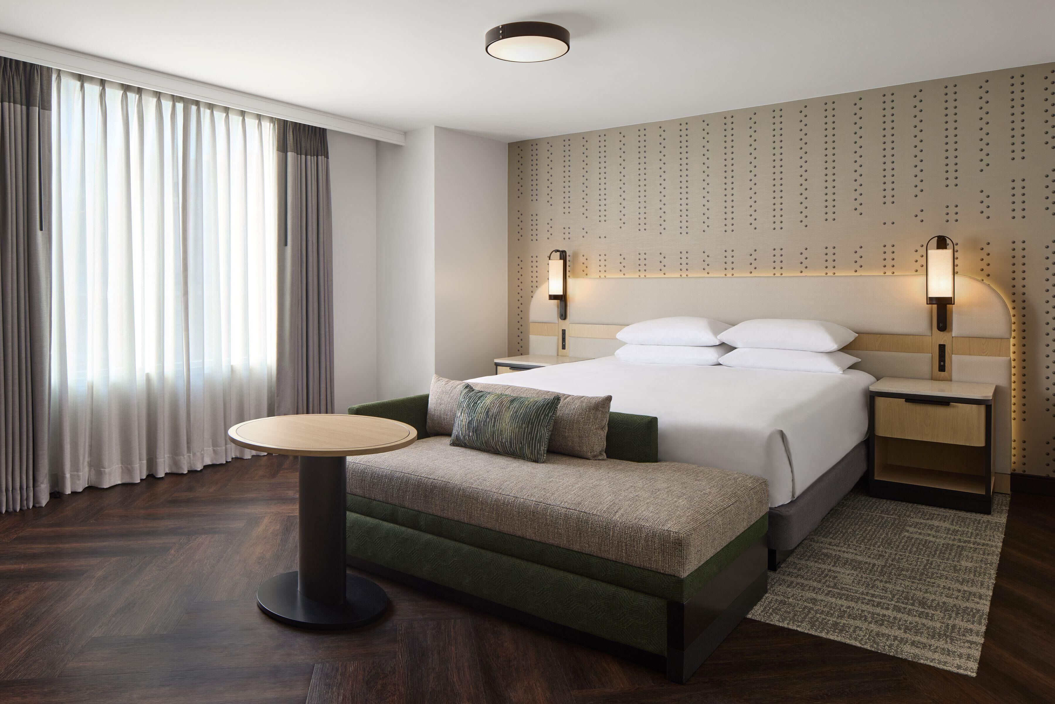 Une chambre d’hôtel moderne, équipée d’un lit, de tables de chevet, d’une table ronde et d’une grande fenêtre avec des rideaux.
