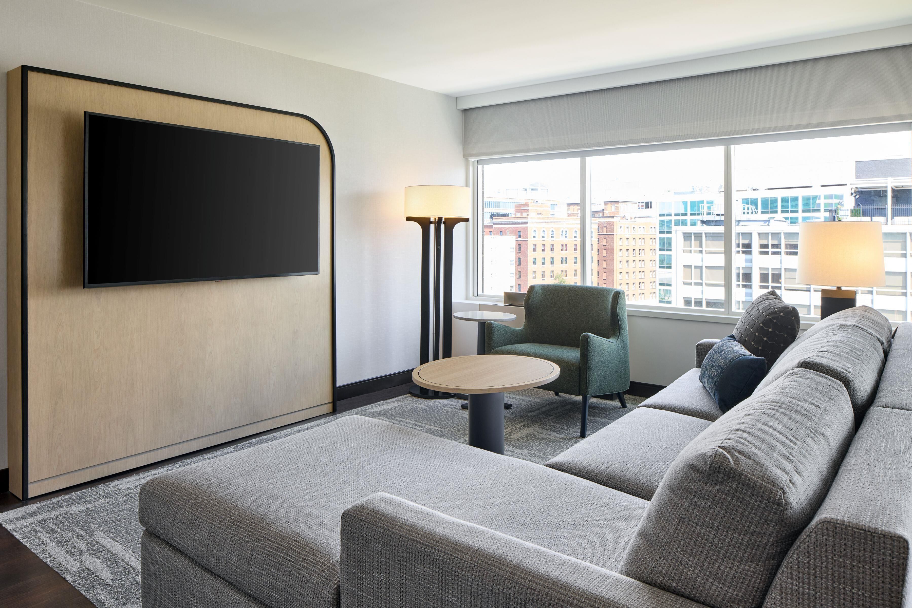 A modern hotel room with a large flat-screen TV mounted on the wall, a sectional sofa, a small round table, a lamp, and a view of the city through large windows.