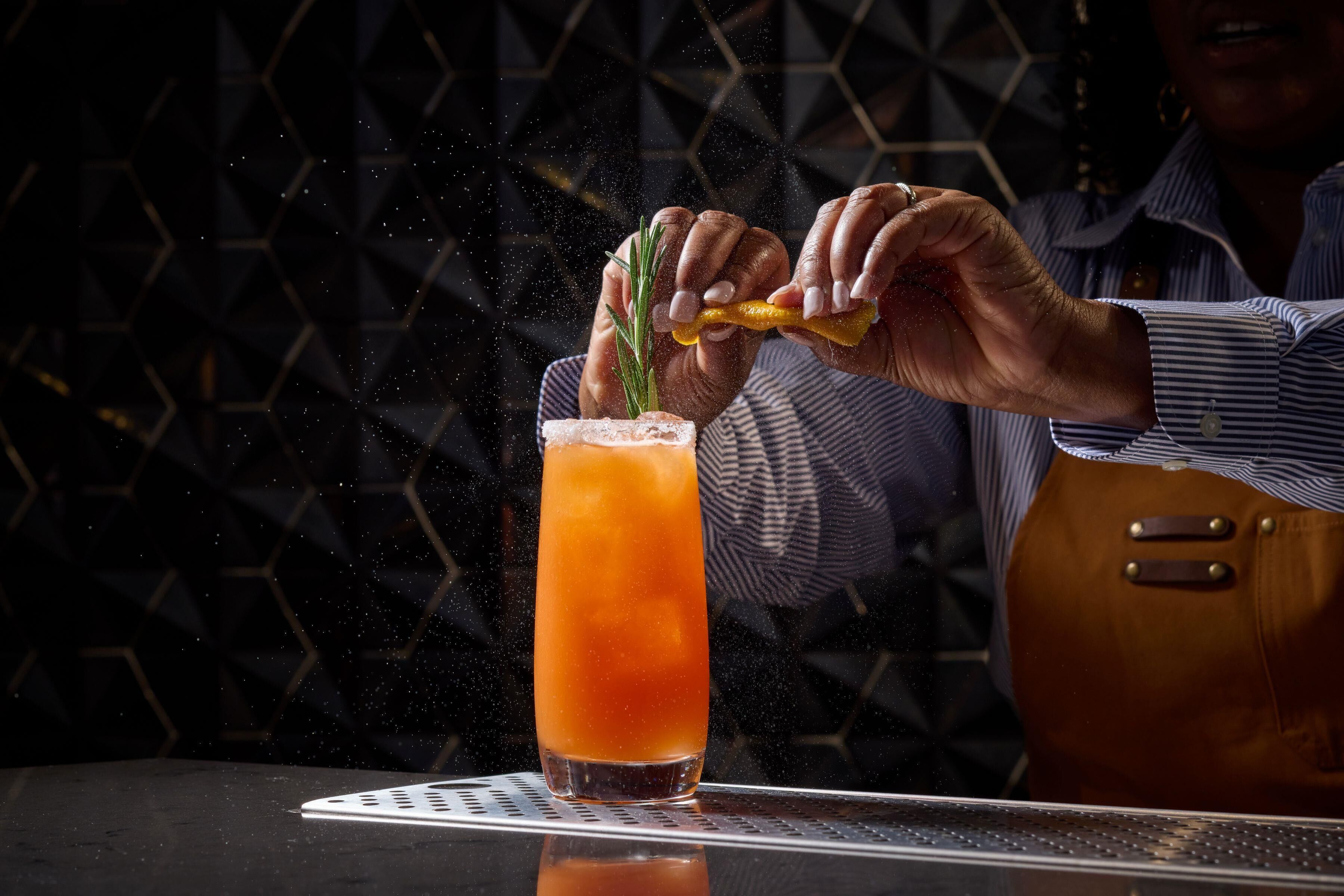 A person pouring a drink into a tall glass with a straw, with a geometric patterned wall in the background.