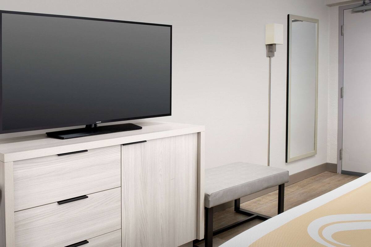 Habitación de hotel moderna con televisión de pantalla plana sobre un mueble de color claro, un banco y un espejo grande en la pared.