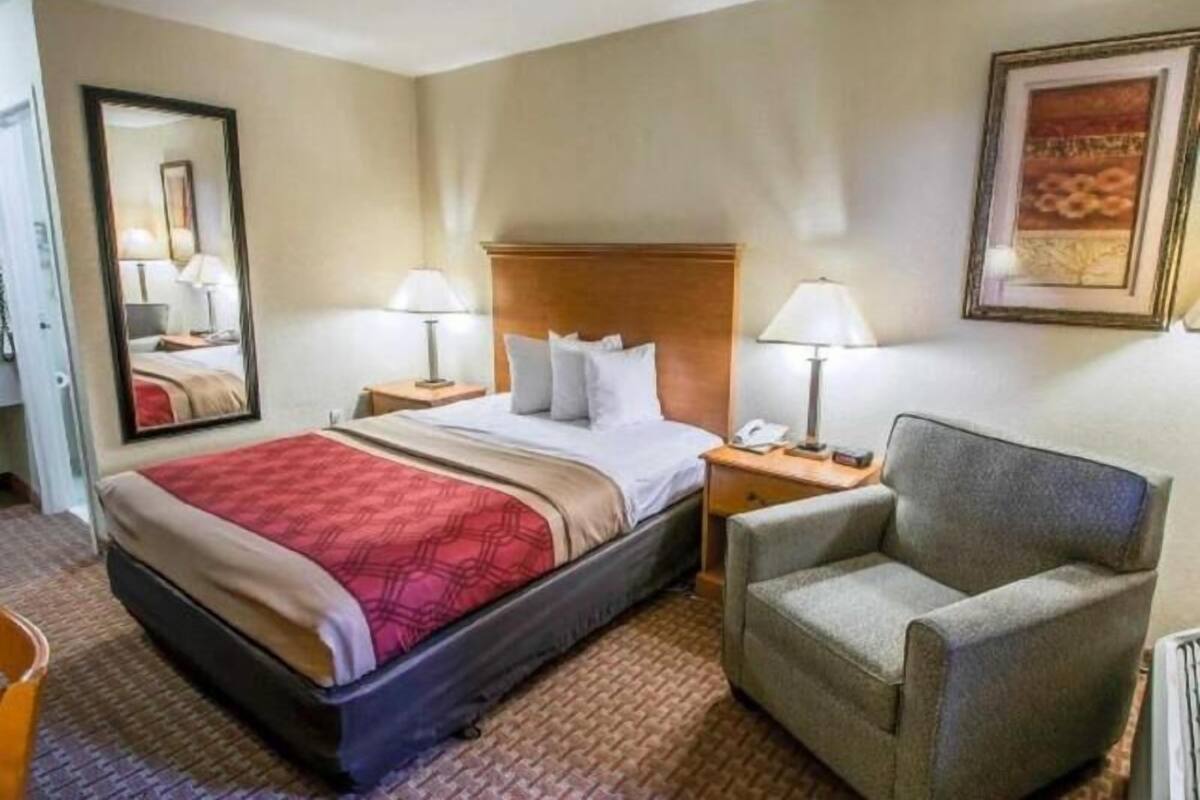 Habitación de hotel con cama, dos mesitas de noche, una silla, un espejo y un cuadro en la pared.