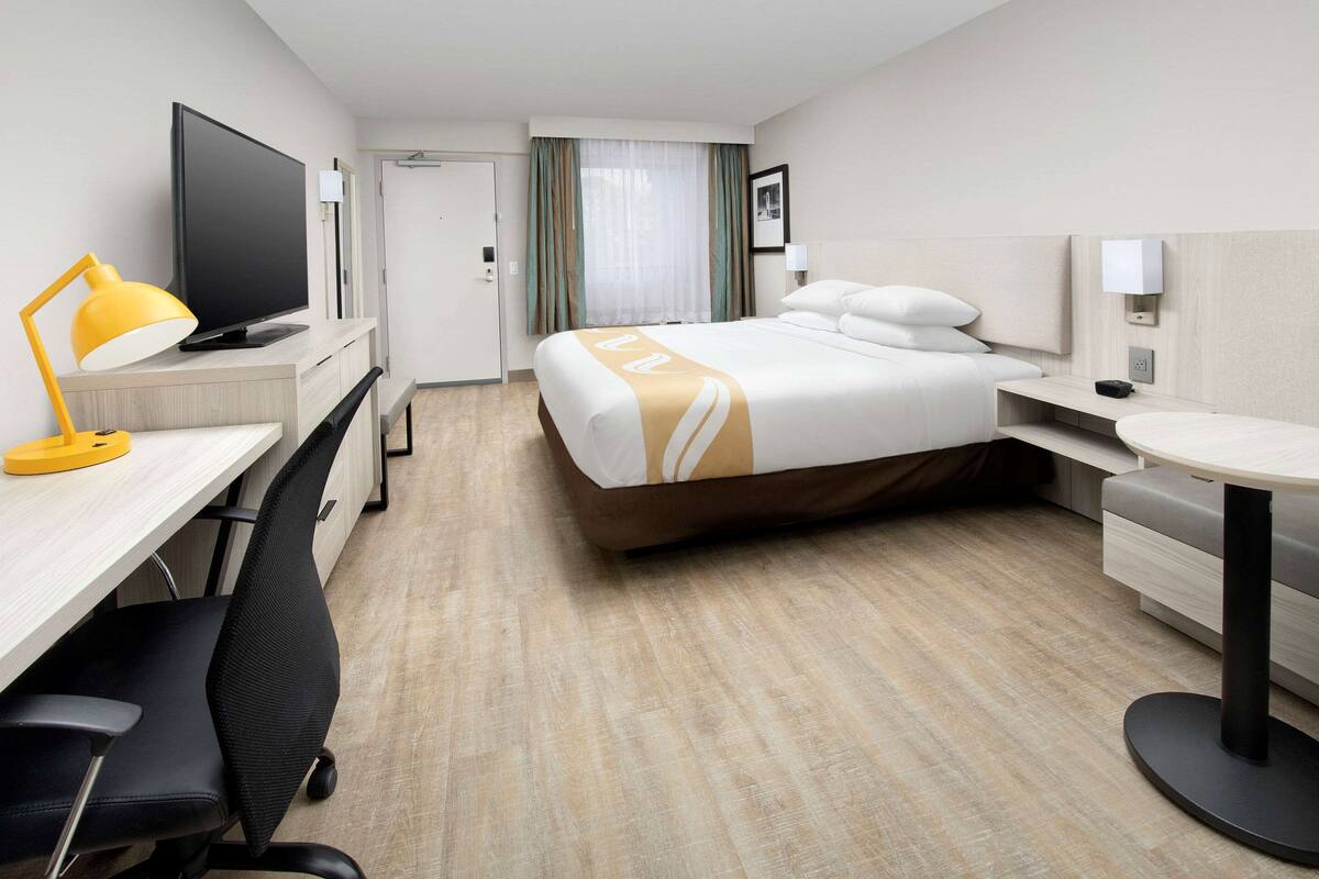 Una habitación de hotel con una cama, un escritorio con computadora, televisor, lámpara, silla y una mesita.