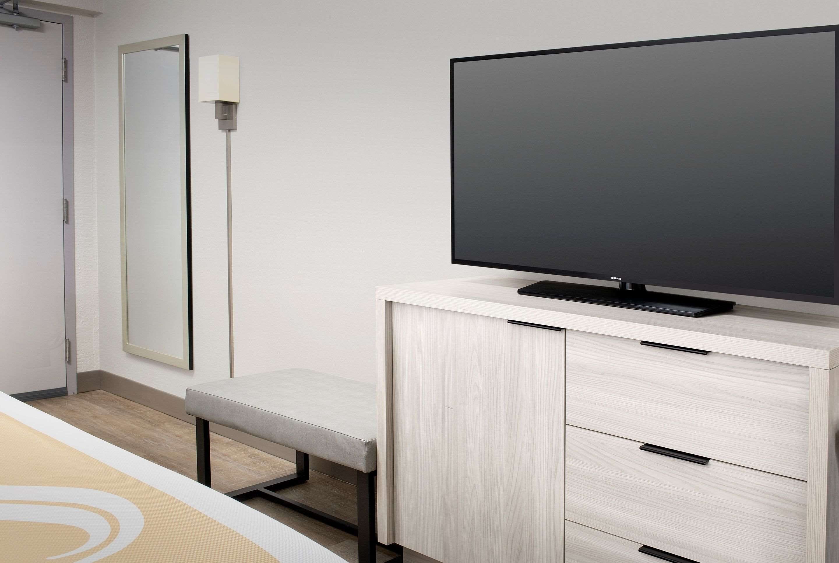 Habitació d'hotel moderna amb un gran televisor de pantalla plana sobre un moble de fusta, una banca i un mirall gran a la paret.