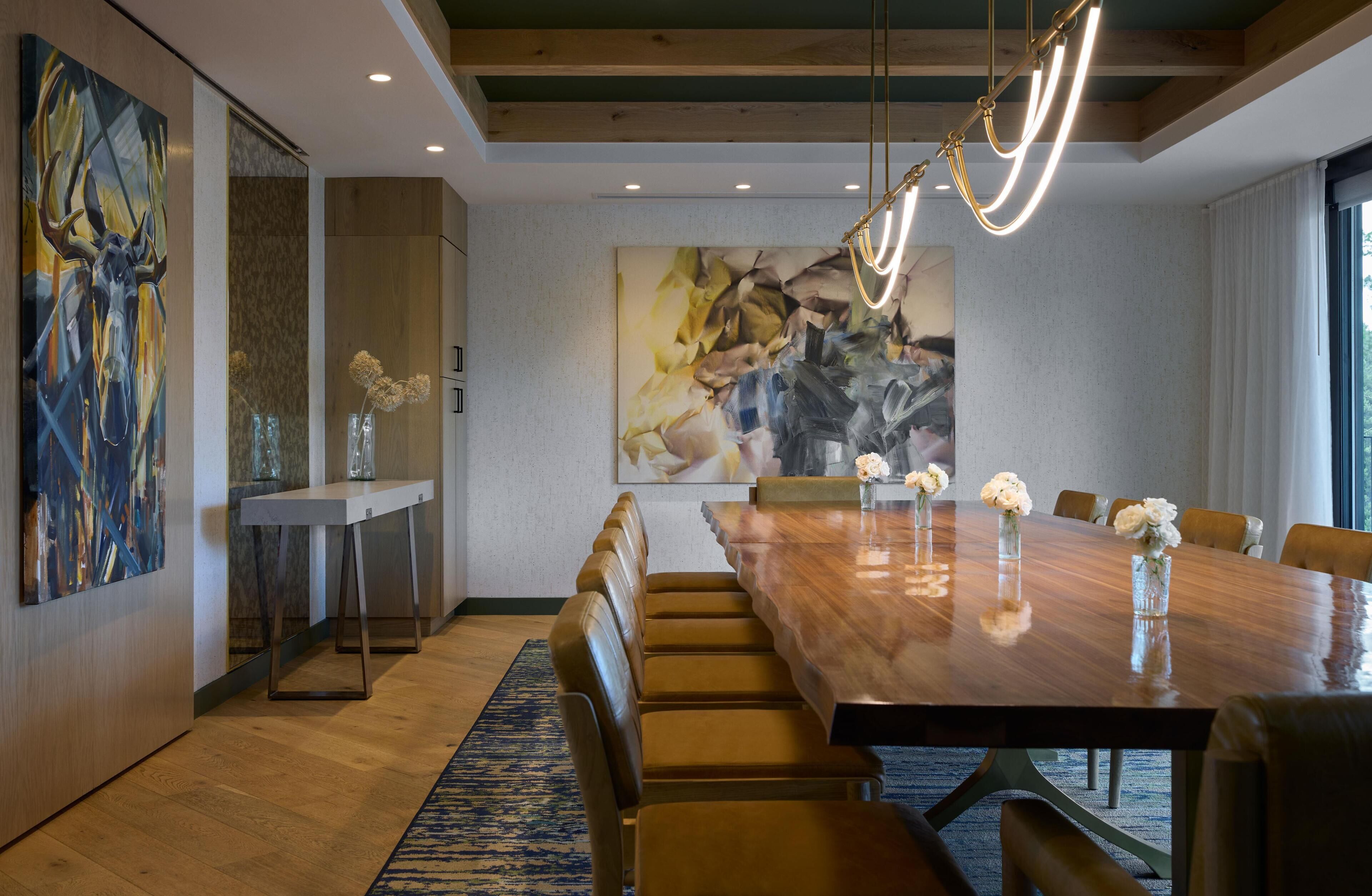 Une salle à manger moderne avec une longue table en bois, des chaises en cuir et des œuvres d’art abstraites accrochées aux murs.