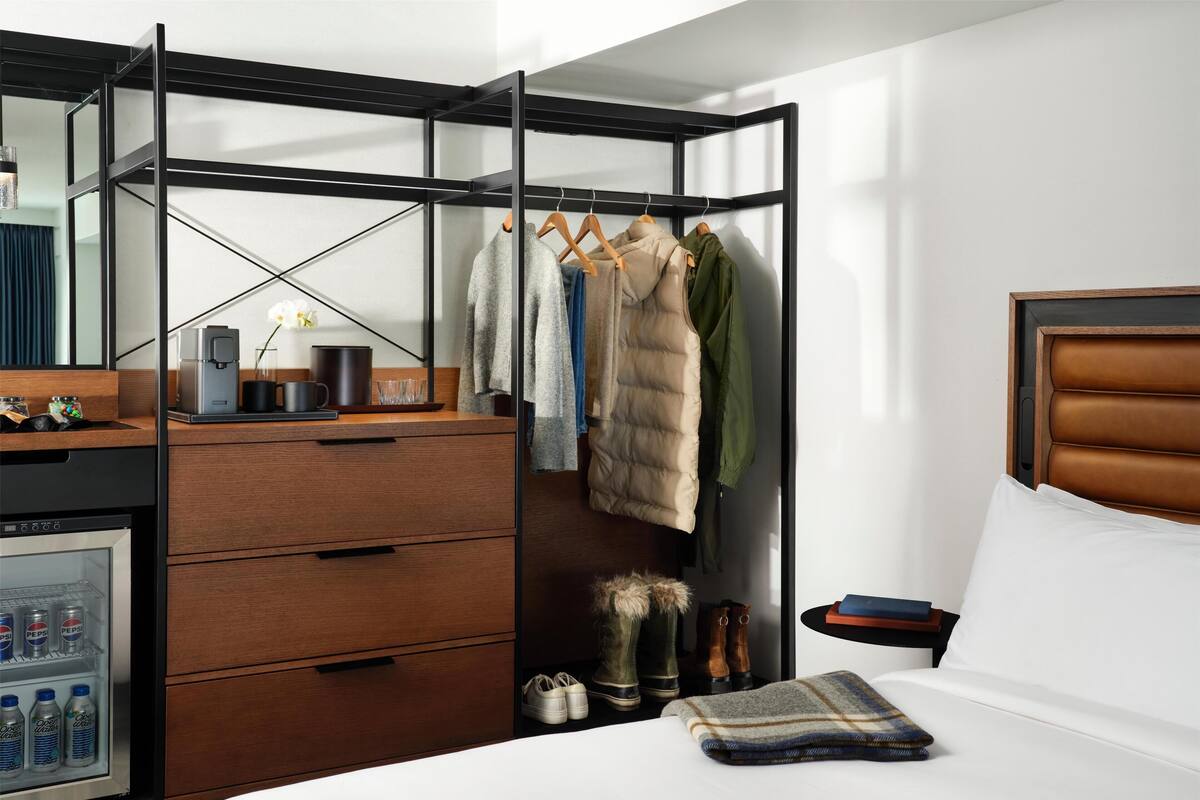 A hotel room with a wardrobe, a bed, a bedside table with a book, a mini-fridge with beverages, and a shoe rack.