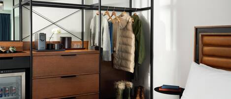 A hotel room with a wardrobe, a bed, a bedside table with a book, a mini-fridge with beverages, and a shoe rack.