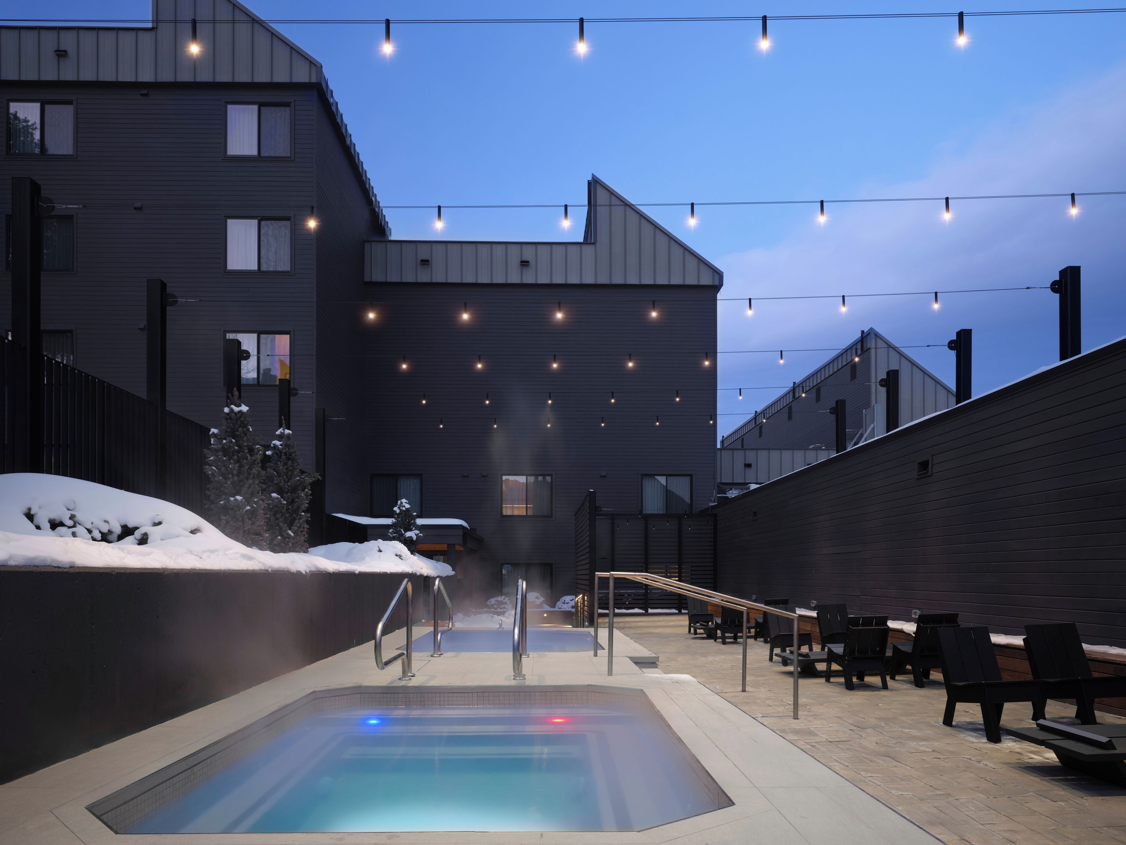 A heated outdoor pool area with a modern building in the background, surrounded by snow-covered ground.
