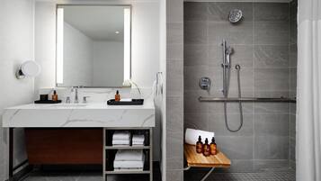 Un baño moderno con tocador de mármol, un espejo grande, ducha con cabezal de ducha de mano y estante con artículos de aseo.