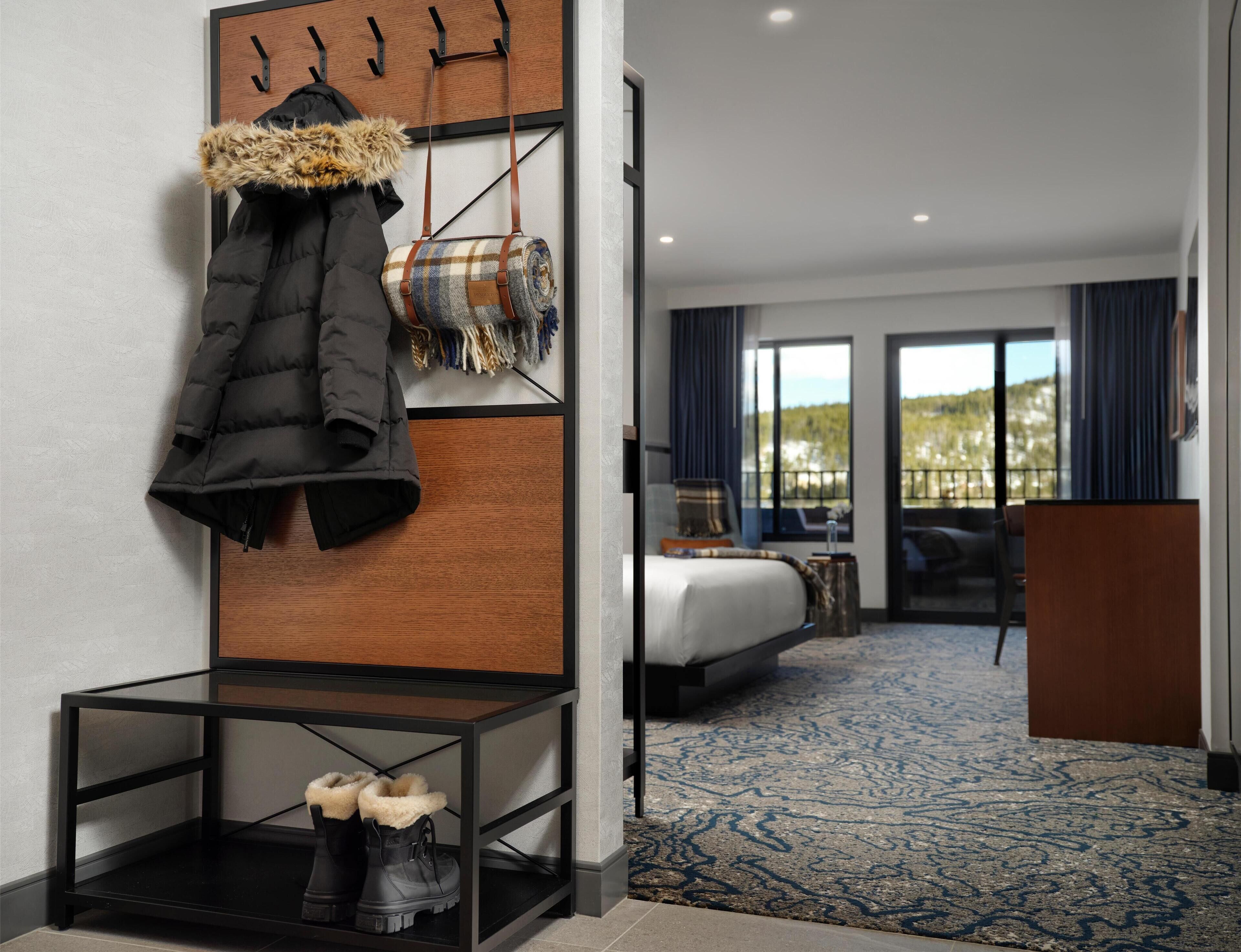 A hotel room with a bed, a seating area with a view, a coat rack, and a table with boots.