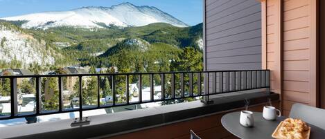 Un balcon avec vue sur une montagne enneigée, une table avec un plateau de nourriture et une chaise.