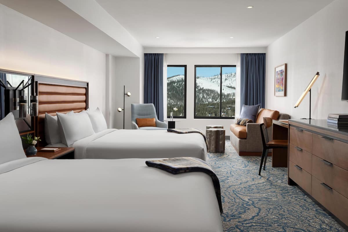 A hotel room with two beds, a desk, a chair, a dresser, and large windows with a mountain view.