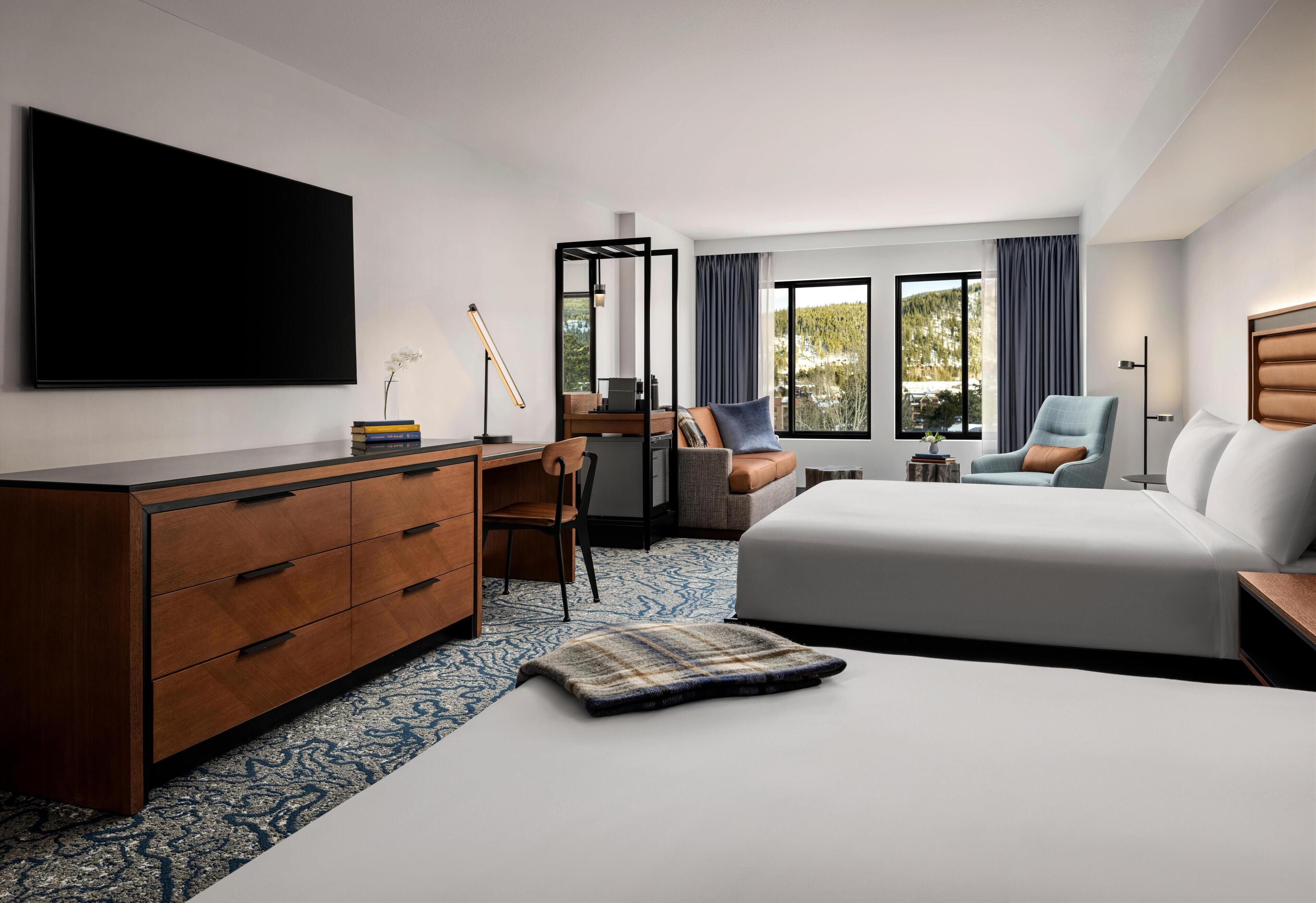 A modern hotel room with a large bed, a desk, a chair, a TV, and a view of the outdoors.