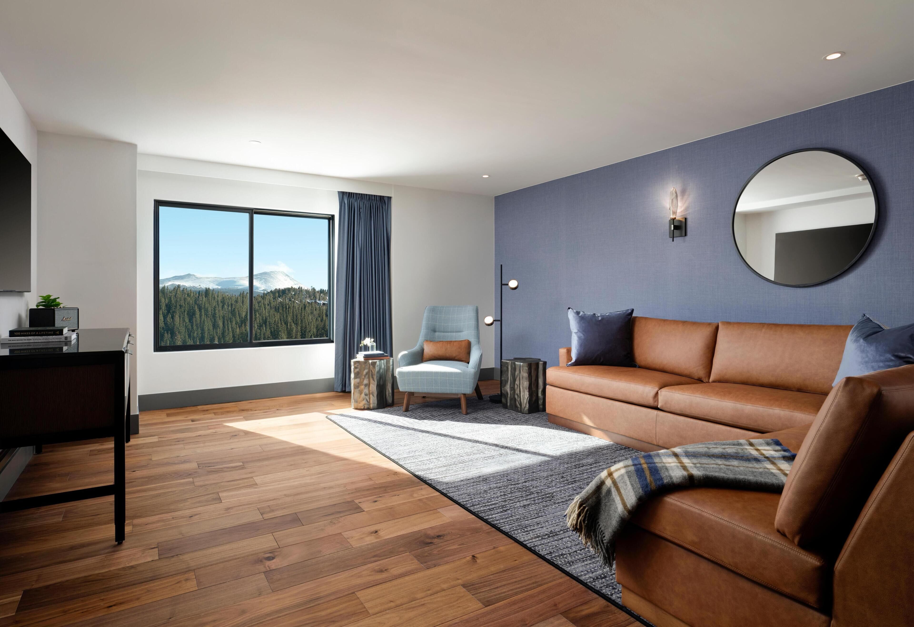 A modern living room with a brown leather sofa, a round mirror, and a large window offering a mountain view.
