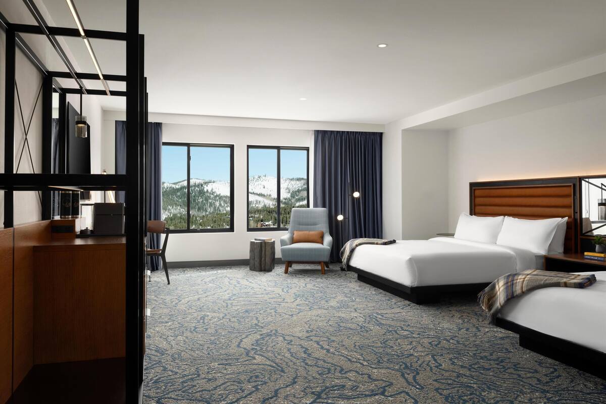 A hotel room with two beds, a desk, a chair, and large windows offering a mountain view.
