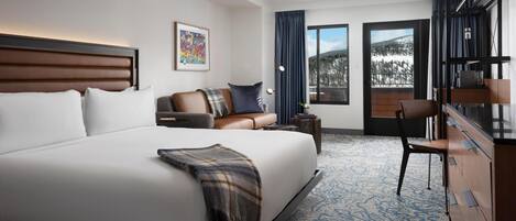 Habitación de hotel moderna con una cama grande, un sofá, un escritorio y vistas a una montaña nevada.