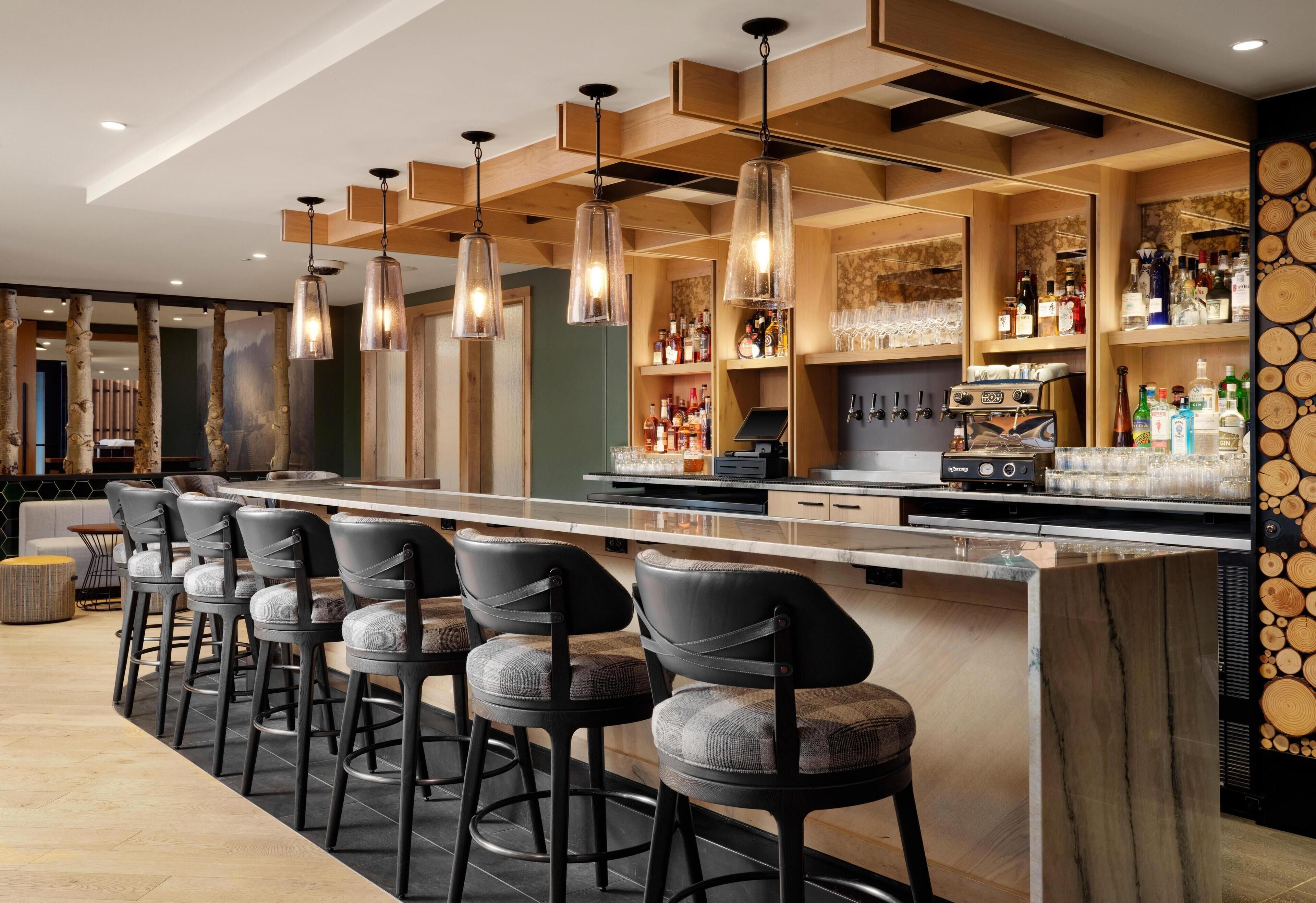 A bar area with a long counter, multiple stools, hanging pendant lights, and shelves stocked with bottles and glasses.