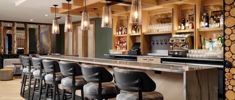 Un espace bar doté d’un long comptoir, de nombreux tabourets, de luminaires suspendus et d’étagères remplies de bouteilles et de verres.