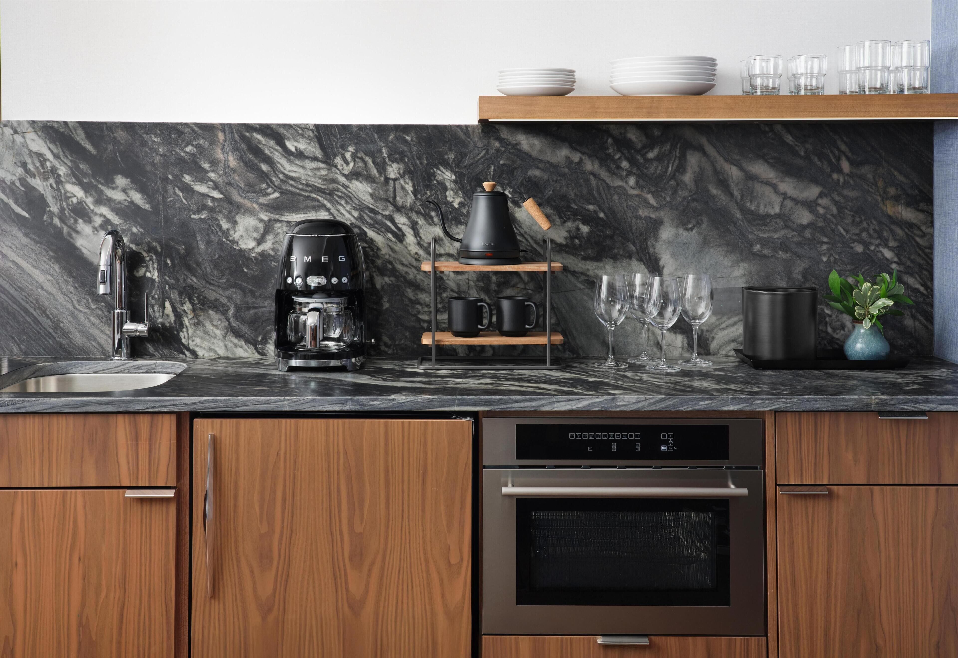 A modern kitchen with a marble backsplash, wooden cabinets, a coffee maker, a kettle, and a built-in oven.