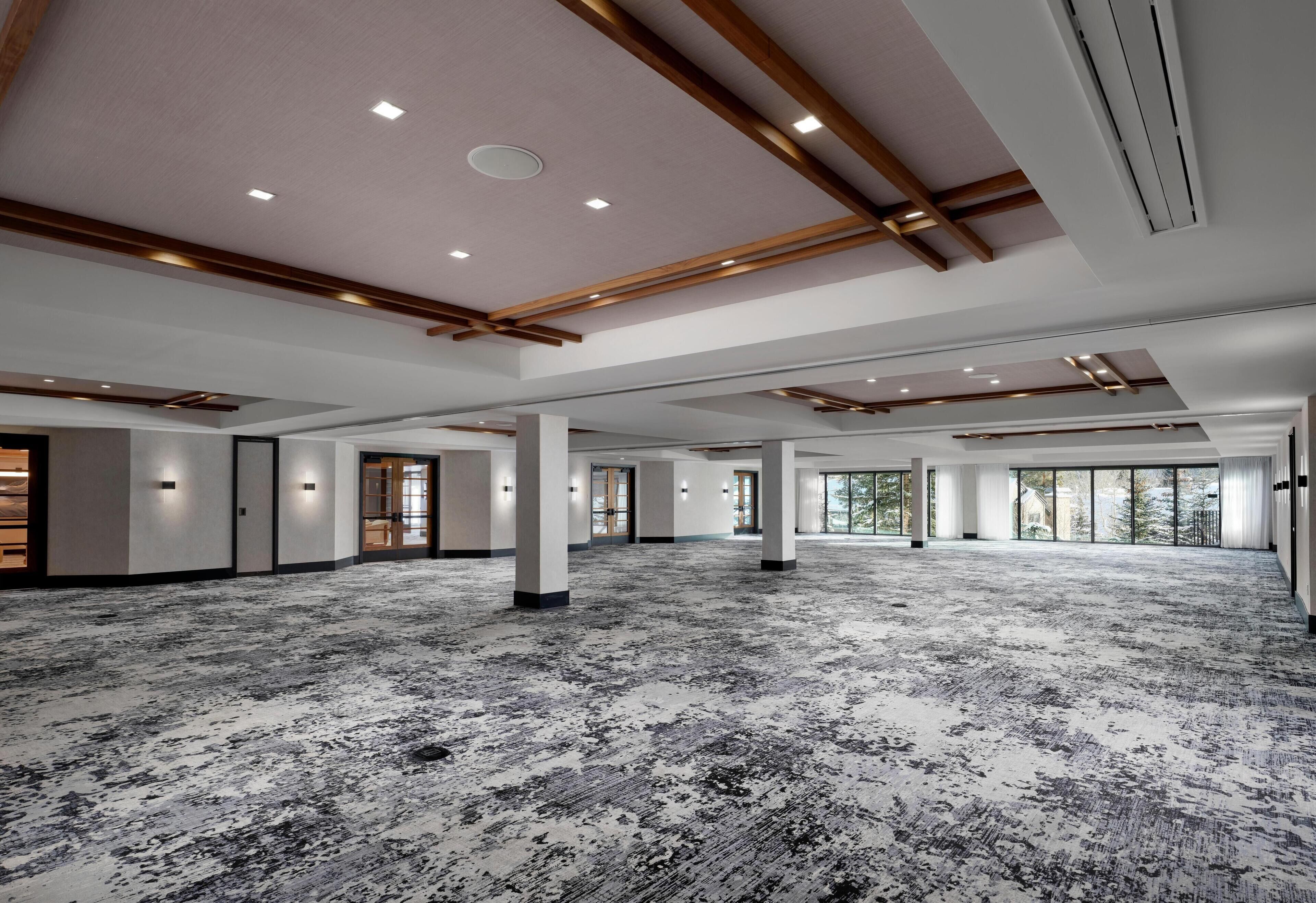 Une salle de conférence spacieuse et bien éclairée, dotée d’un tapis à motifs, de poutres en bois au plafond et de grandes fenêtres laissant entrer la lumière naturelle.