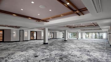 Una espaciosa sala de conferencias, bien iluminada, con alfombra estampada, vigas de madera en el techo y ventanas amplias que permiten la entrada de luz natural.