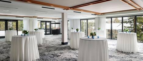 A spacious room with round tables covered in white tablecloths, large windows offering a view of the outdoors, and a carpeted floor.