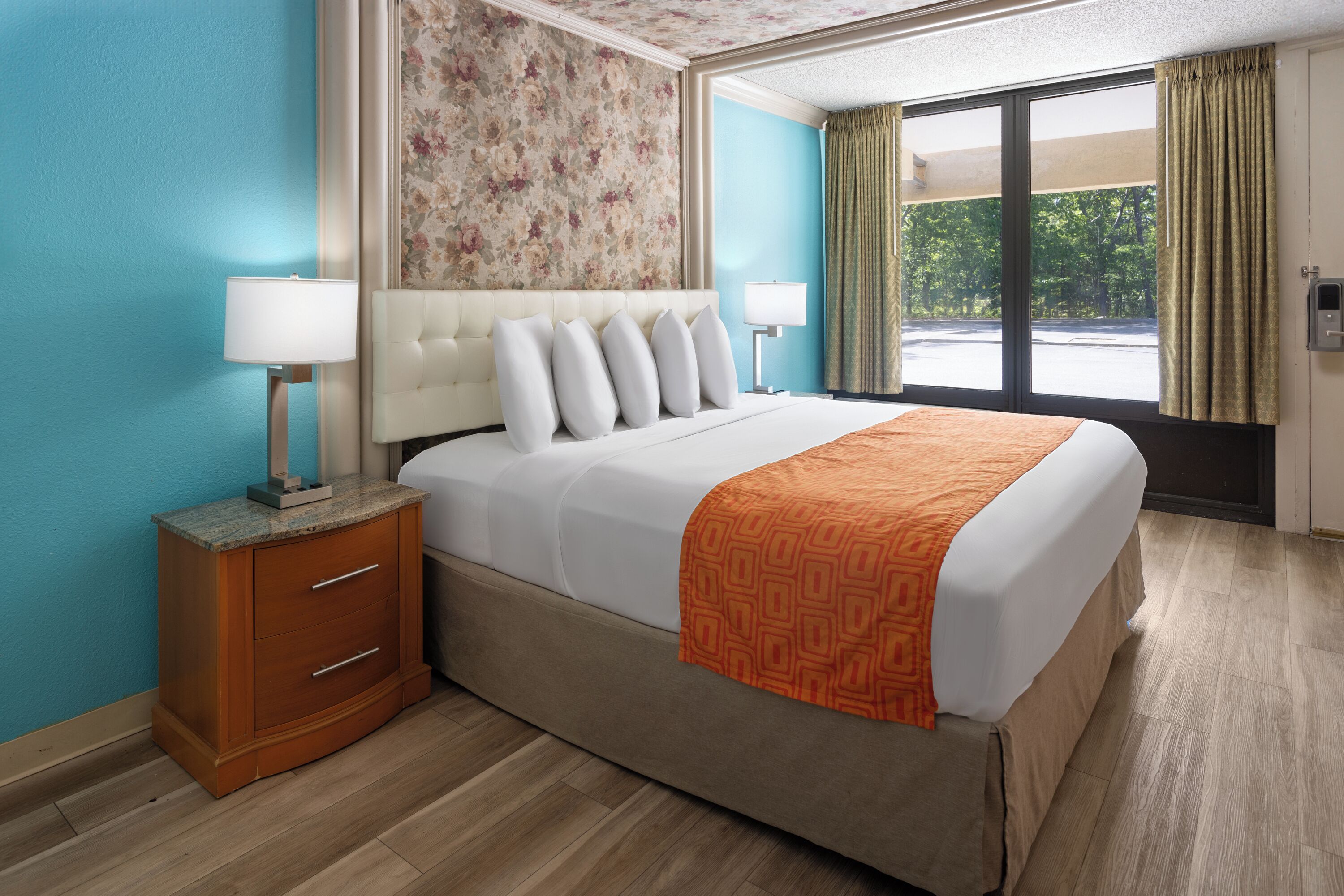 A hotel room with a large bed, two bedside lamps, a wooden nightstand, a floral-patterned headboard, and a view of the outdoors through a window with curtains.