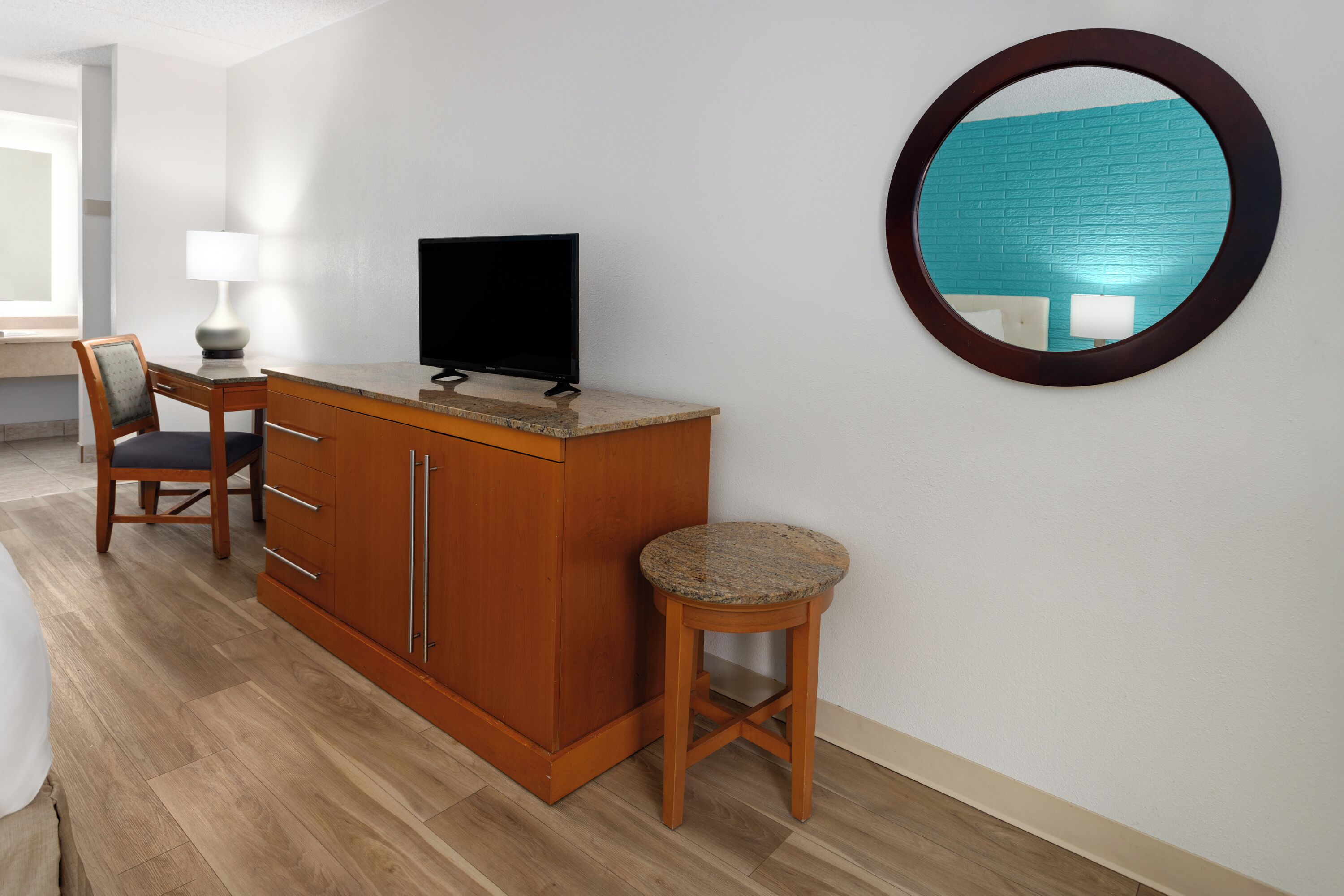 Une chambre d’hôtel avec un bureau en bois, une télévision, un miroir rond et un petit tabouret.