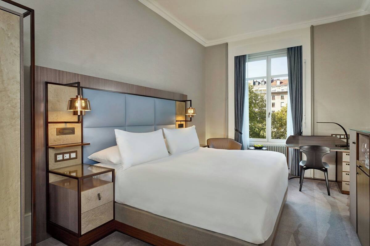 A hotel room with a large bed, a desk, and a chair. There is a window with a view of buildings outside.