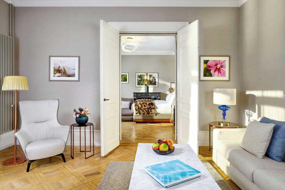 A modern living room with a white armchair, a coffee table with a book, a sofa, and a dining table with fruit.