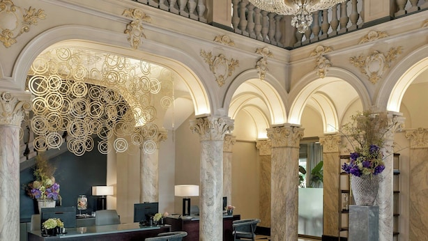 A grand lobby with a checkered floor, ornate columns, a central chandelier, and a reception desk with seating.