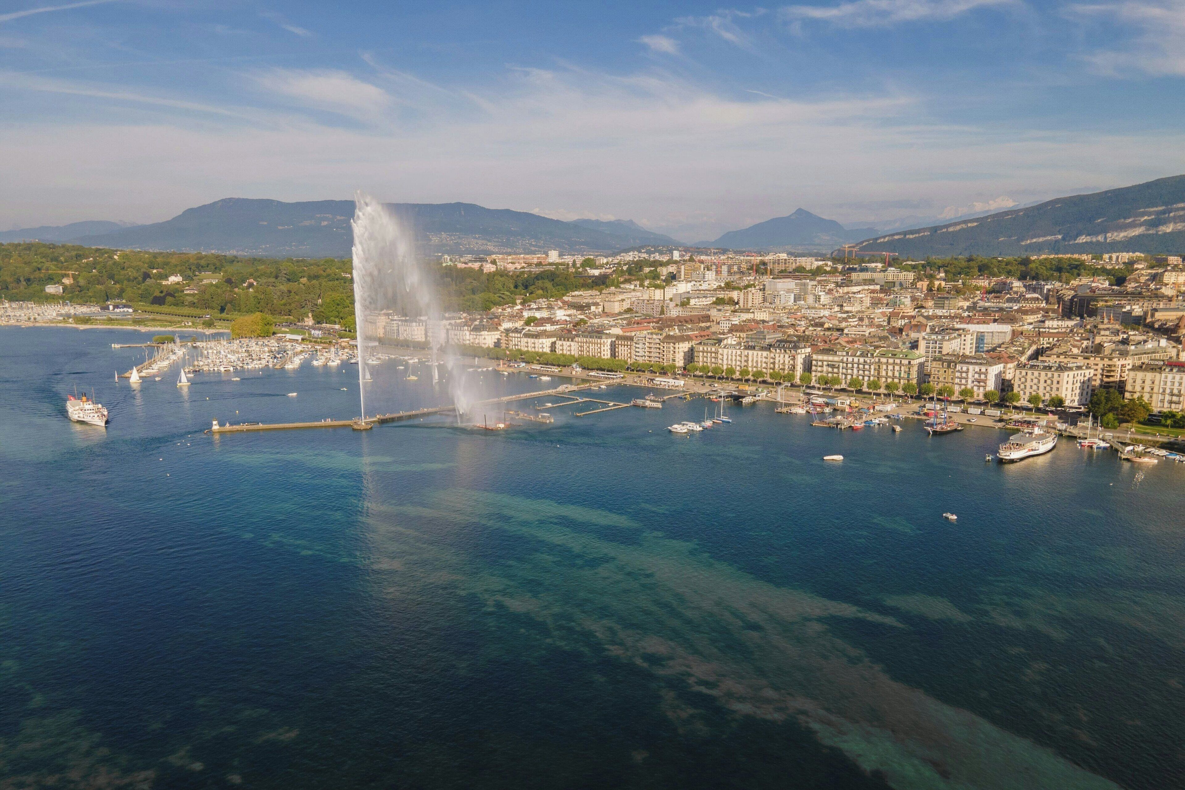 An aerial view of a coastal city with a prominent fountain, marina, and surrounding buildings.